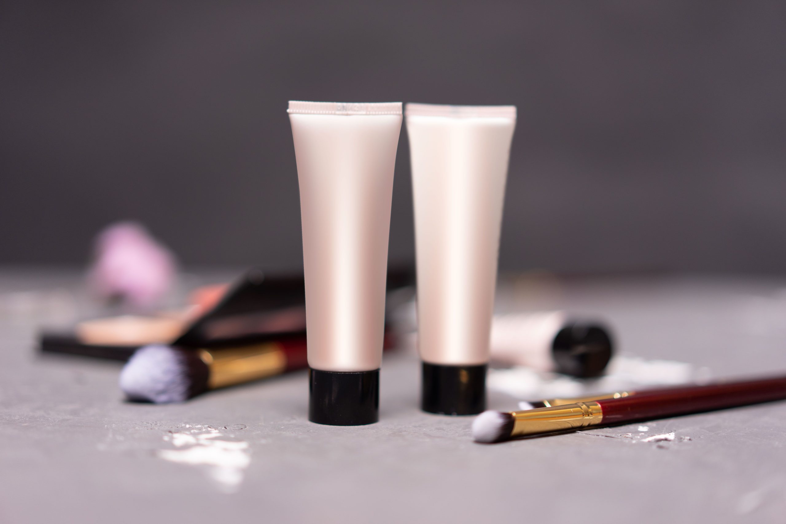 Deux tubes de crème cosmétiques sur fond gris, entourés de pinceaux et de traces de produit, illustrant l